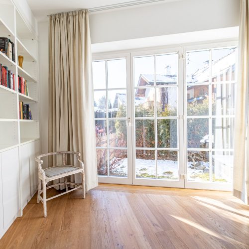 Fensterfront mit creme farbenen Vorhängen und einem kleinen Stuhl in Ferienwohnung in Garmisch-Partenkirchen
